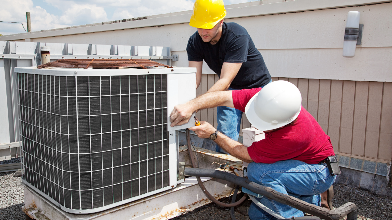Two men are working on an air conditioner on the roof of a building.