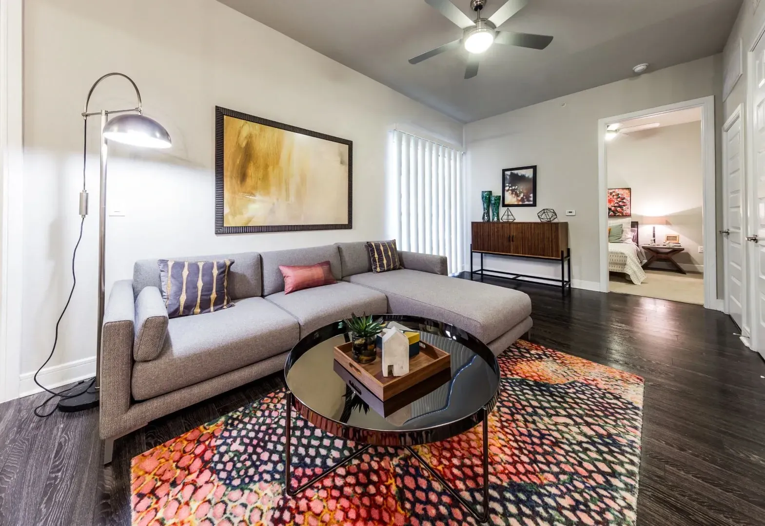 Living room with a sectional sofa, coffee table, and accent rug. A bedroom is visible through an open doorway.