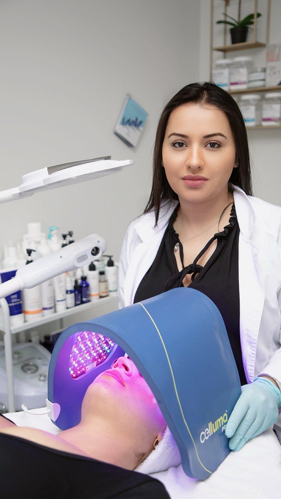 Woman with a client undergoing light therapy in a spa; another woman in a lab coat and gloves looks on.