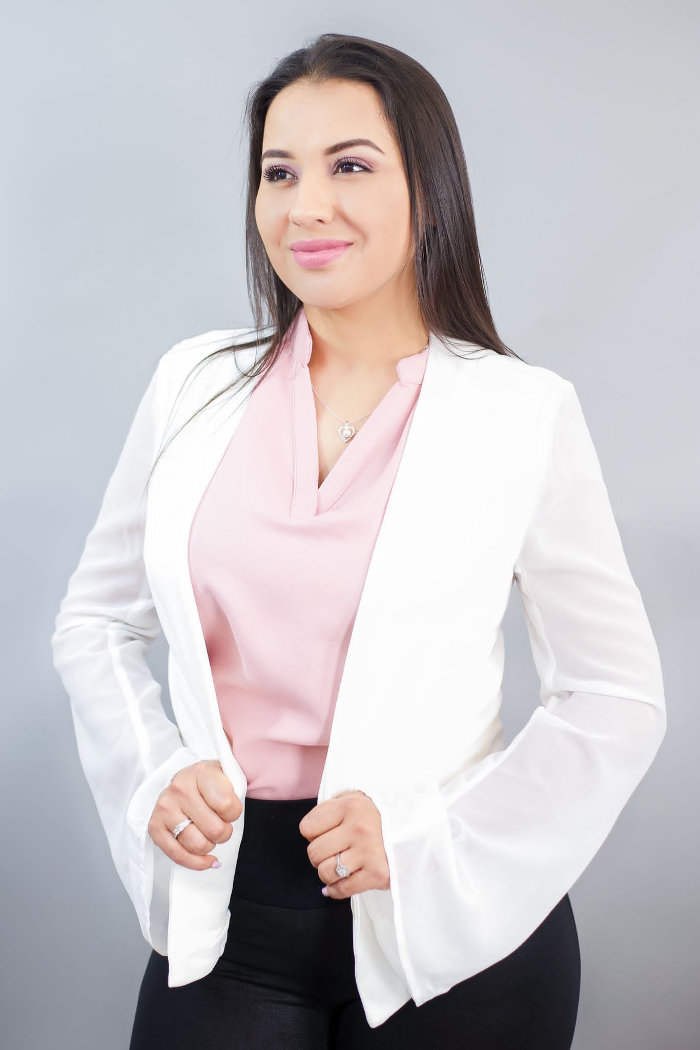 Woman in white blazer and pink top, holding the blazer open, smiling, against a gray background.