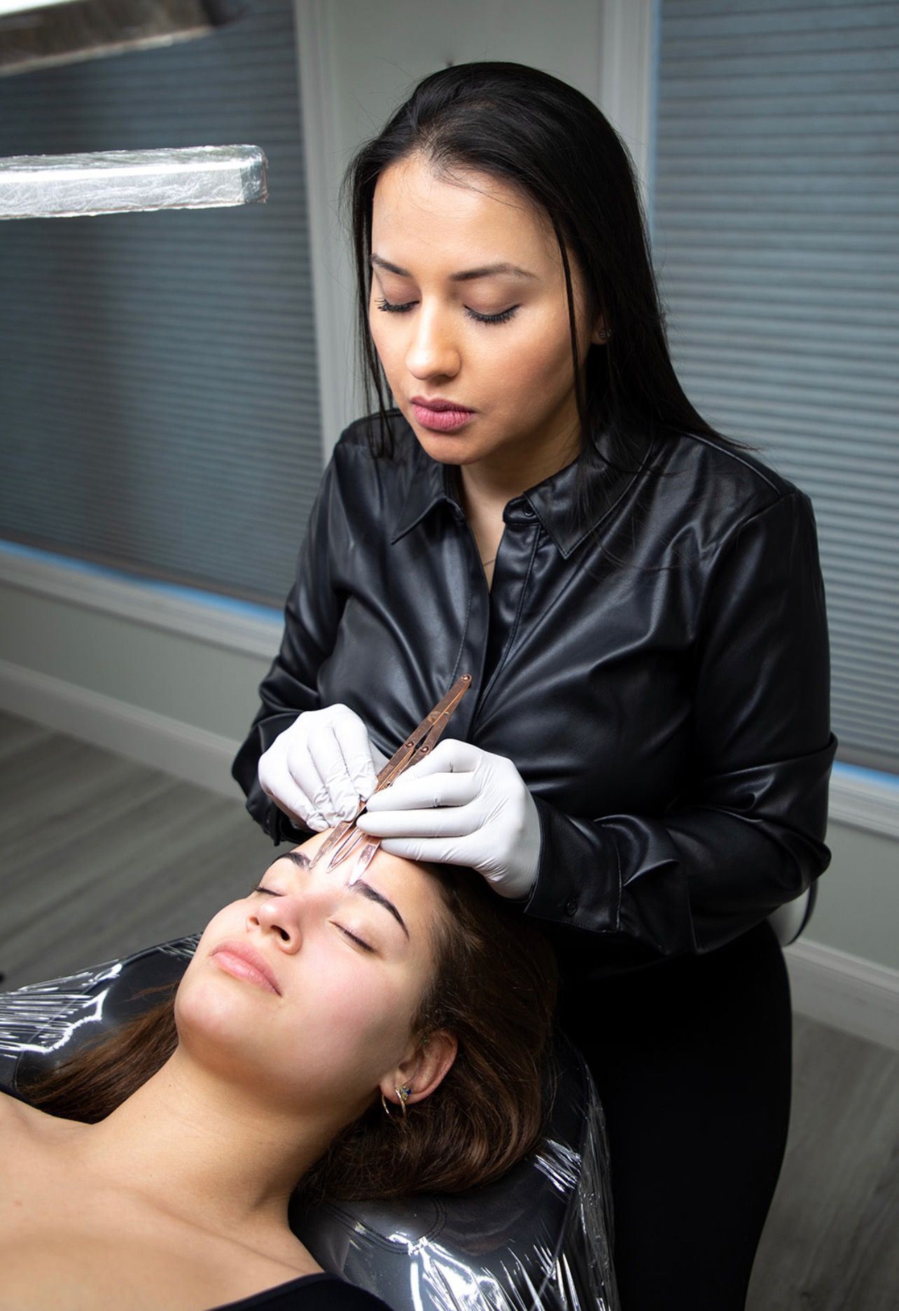 Woman in gloves shaping eyebrows of a client, indoors. Both have dark hair.