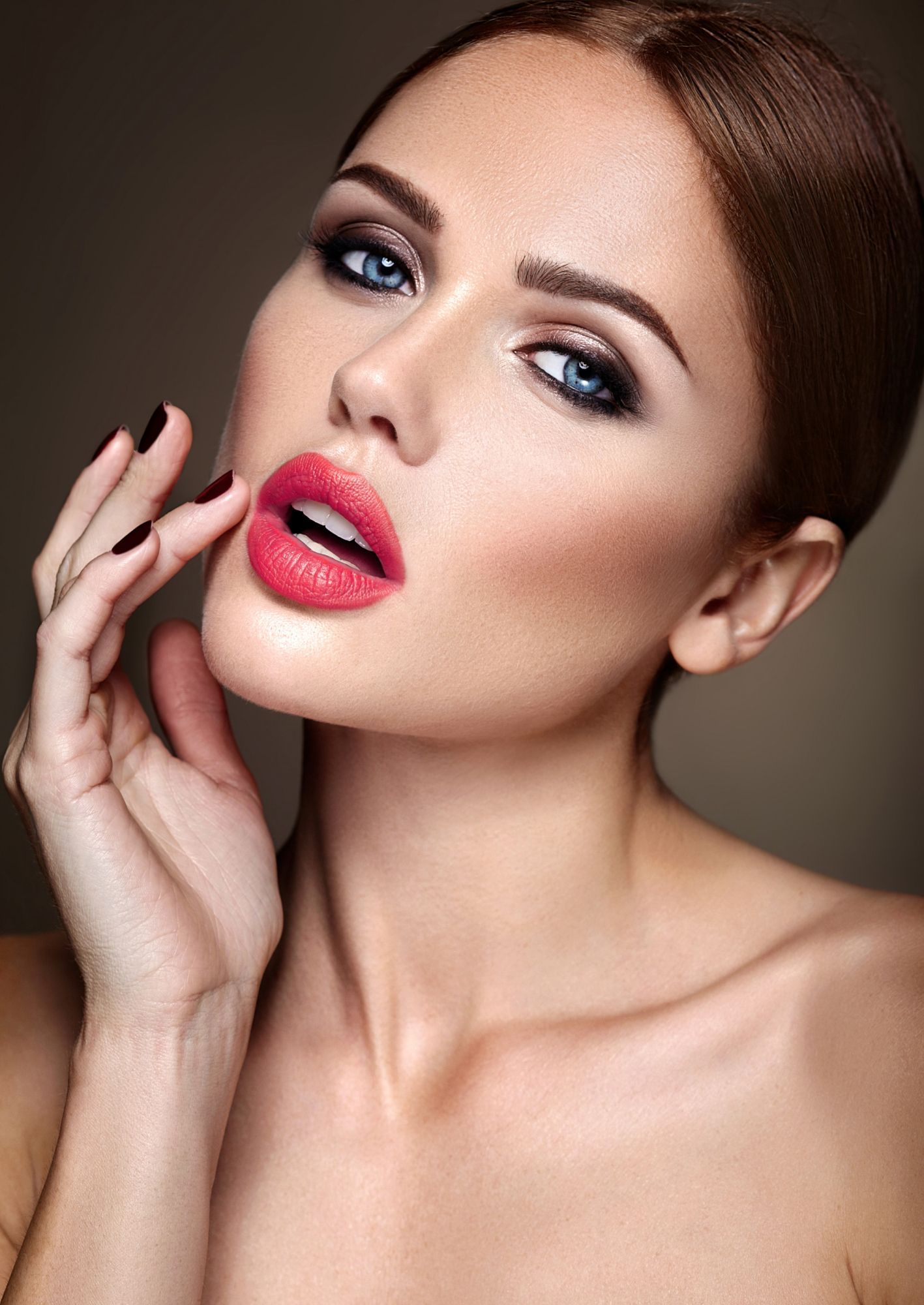 Woman with red lipstick, dramatic eye makeup, and brown hair, looking forward and touching her cheek.