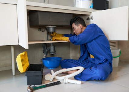 A plumber wearing a blue uniform and yellow gloves repairs a sink pipe while sitting on the floor with tools nearby.