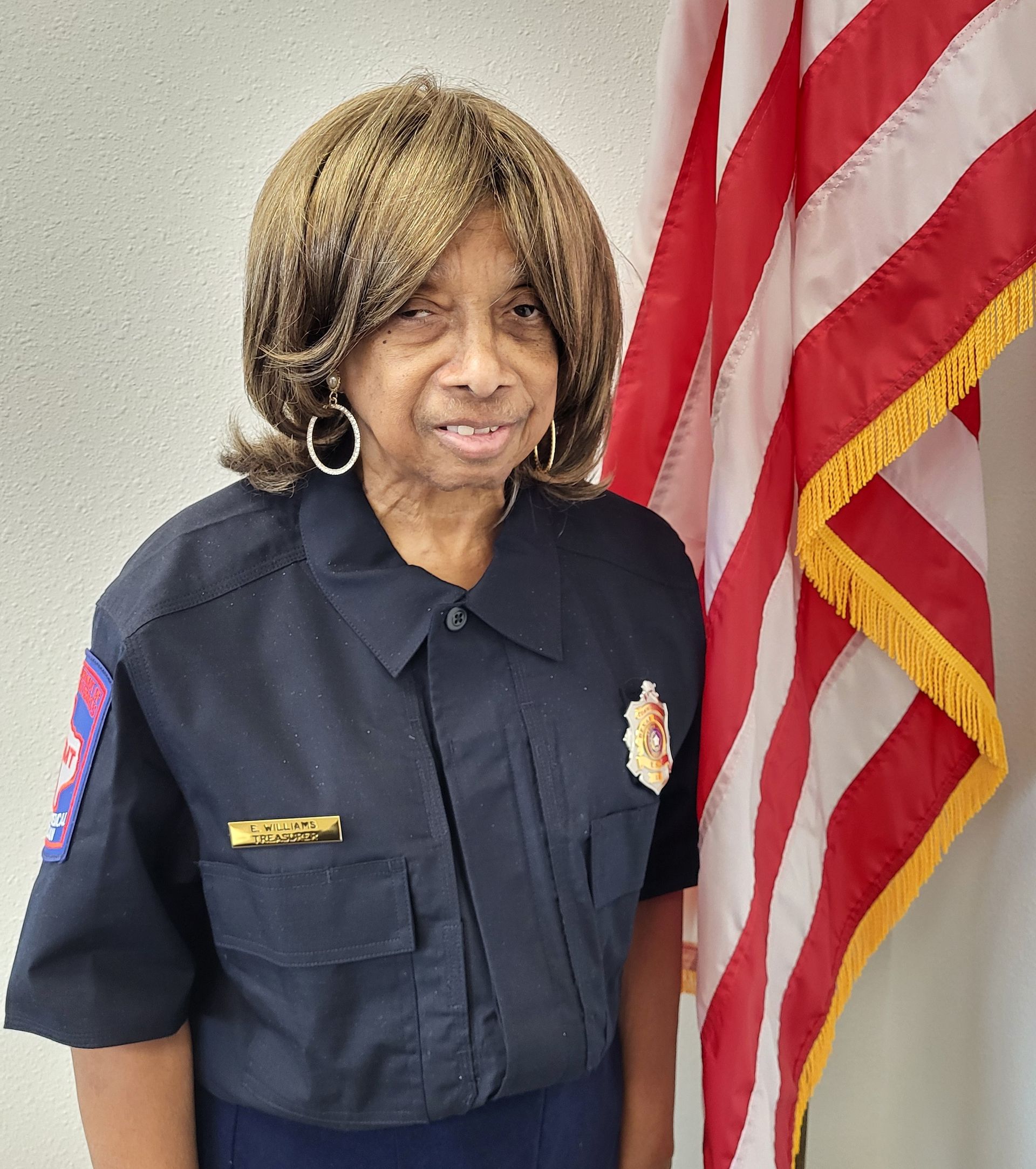 A woman wearing glasses and a blue shirt is standing in front of an American flag.