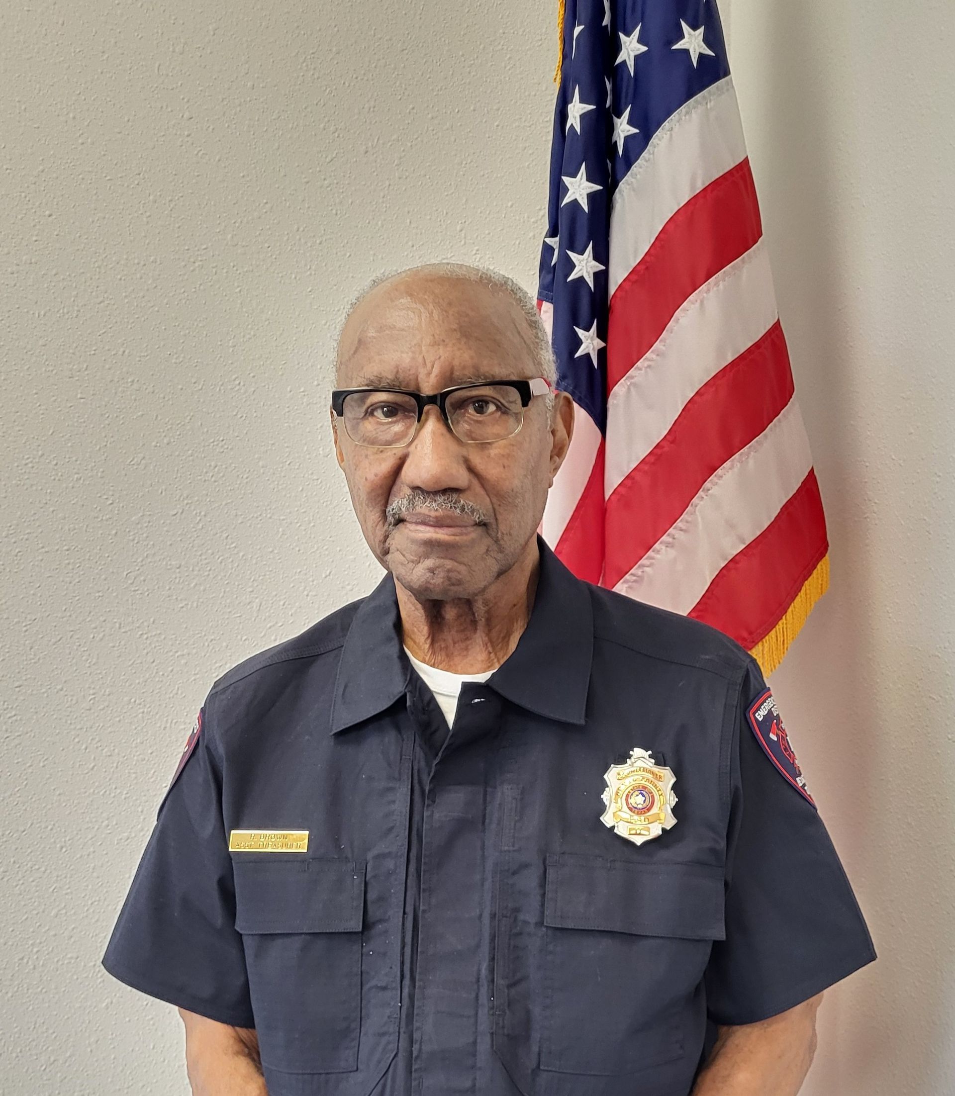 A man wearing glasses is standing in front of an American flag.