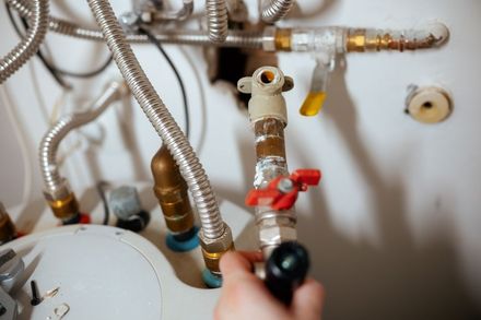 A hand adjusts a valve on water heater plumbing pipes, with braided steel supply lines visible against a white wall.