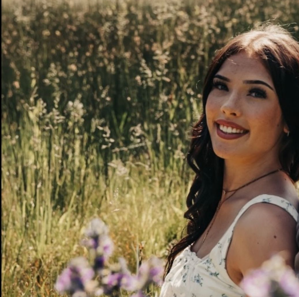 Woman smiles, sitting in field of wildflowers, wearing white floral top.
