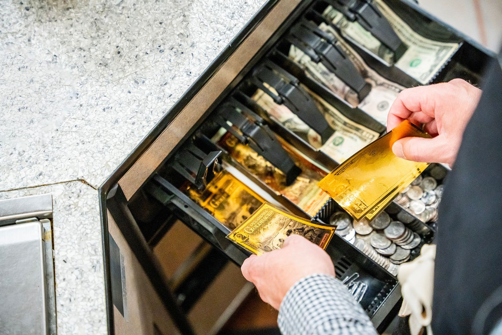 Hands reaching into a cash register drawer holding gold-colored items, with currency and coins visible.