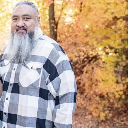 Man with long gray beard in black and white plaid shirt, autumn foliage background.