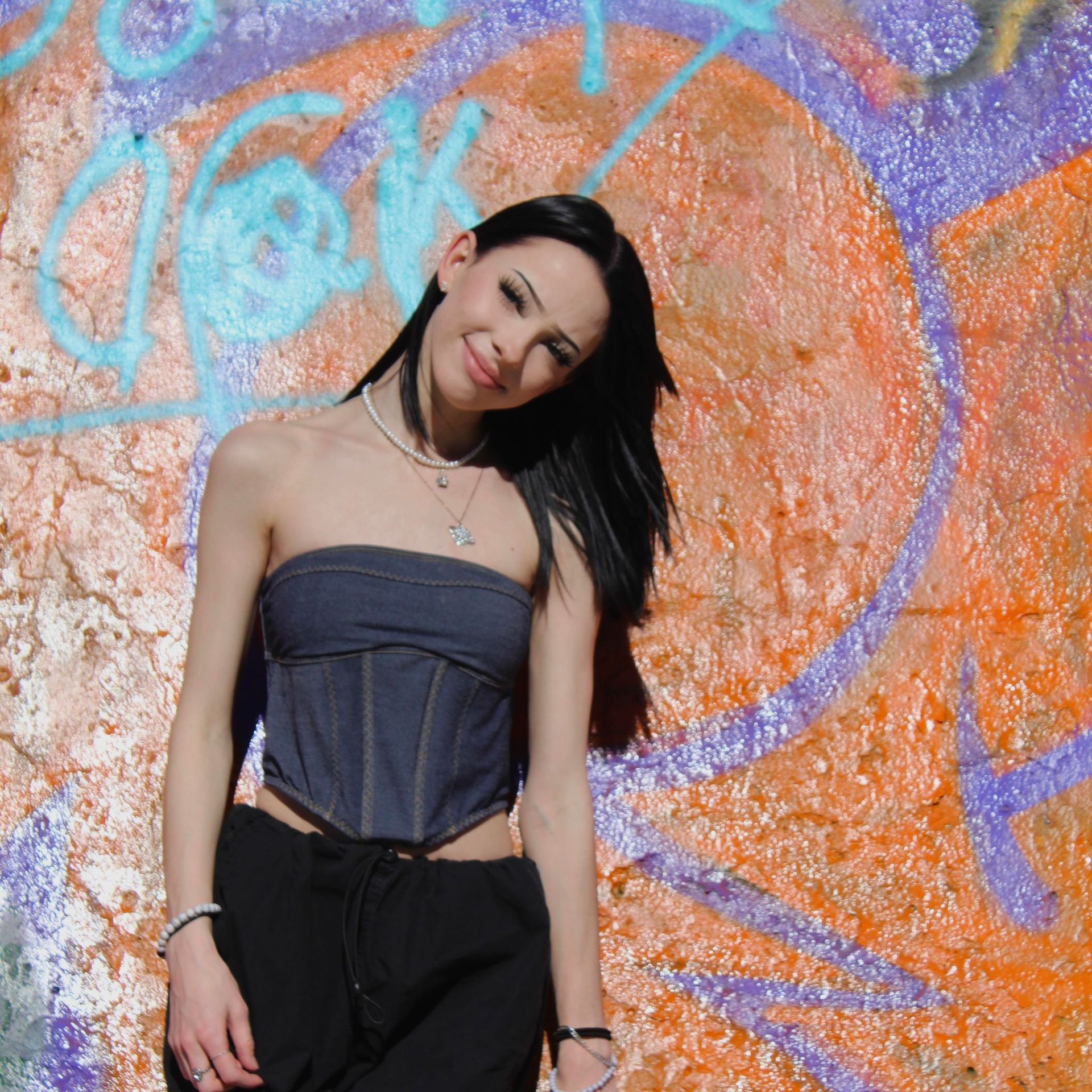 Woman in denim top and black pants poses by graffiti wall.