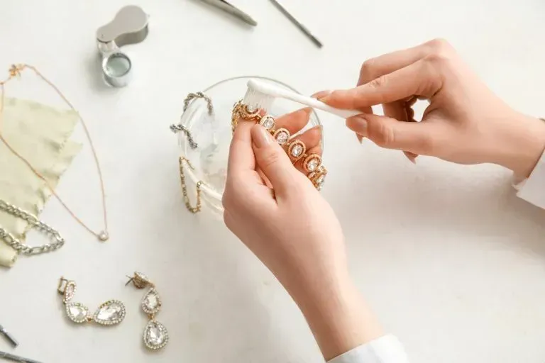 Person cleaning gold jewelry with a cotton swab in a small glass bowl on a white surface.