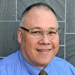 Man holding a fish outdoors, wearing a black shirt and cap. The fish is greenish-brown. Green background.