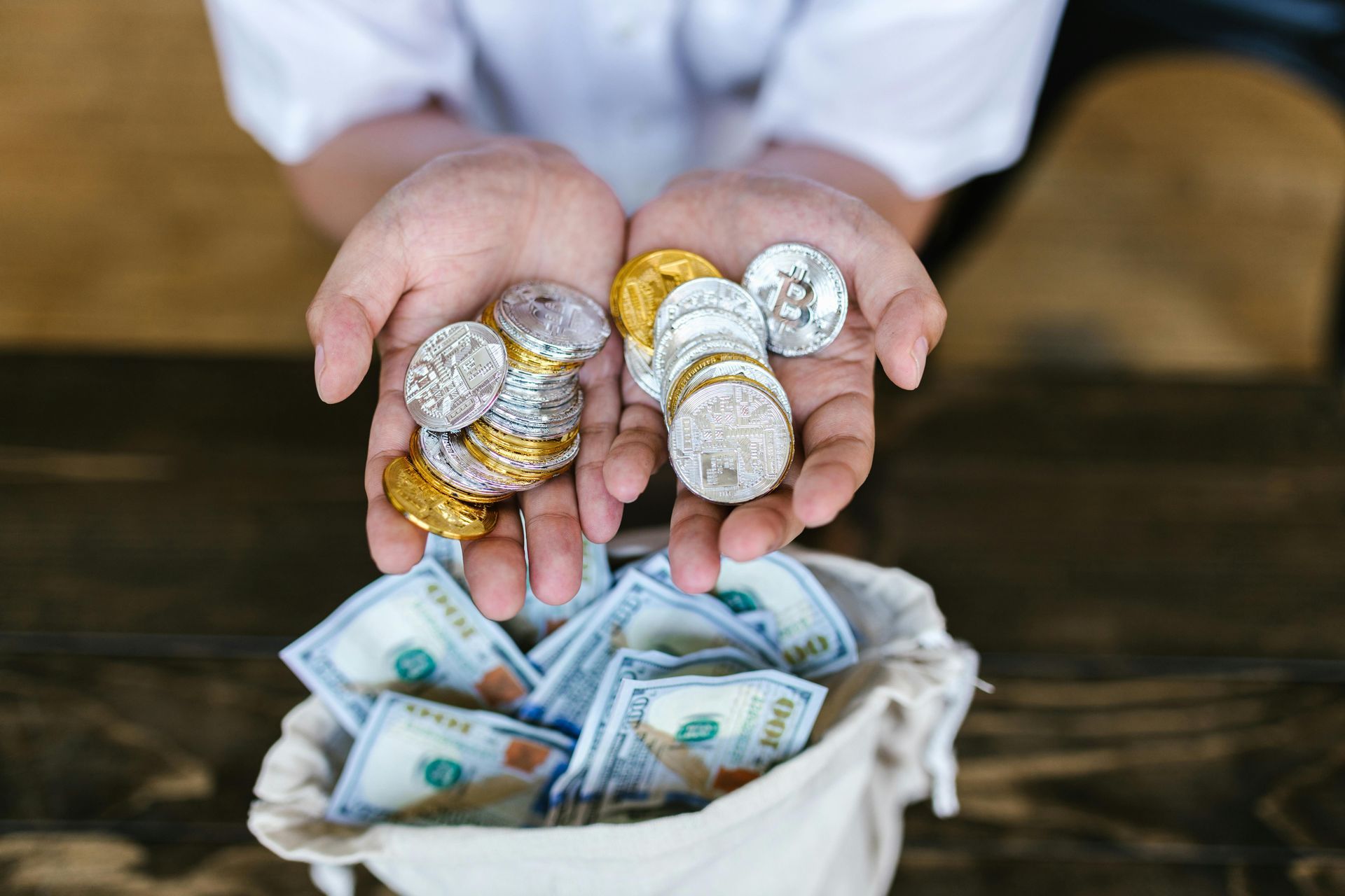 Hands holding coins above a bag overflowing with US dollar bills, on a wooden surface.