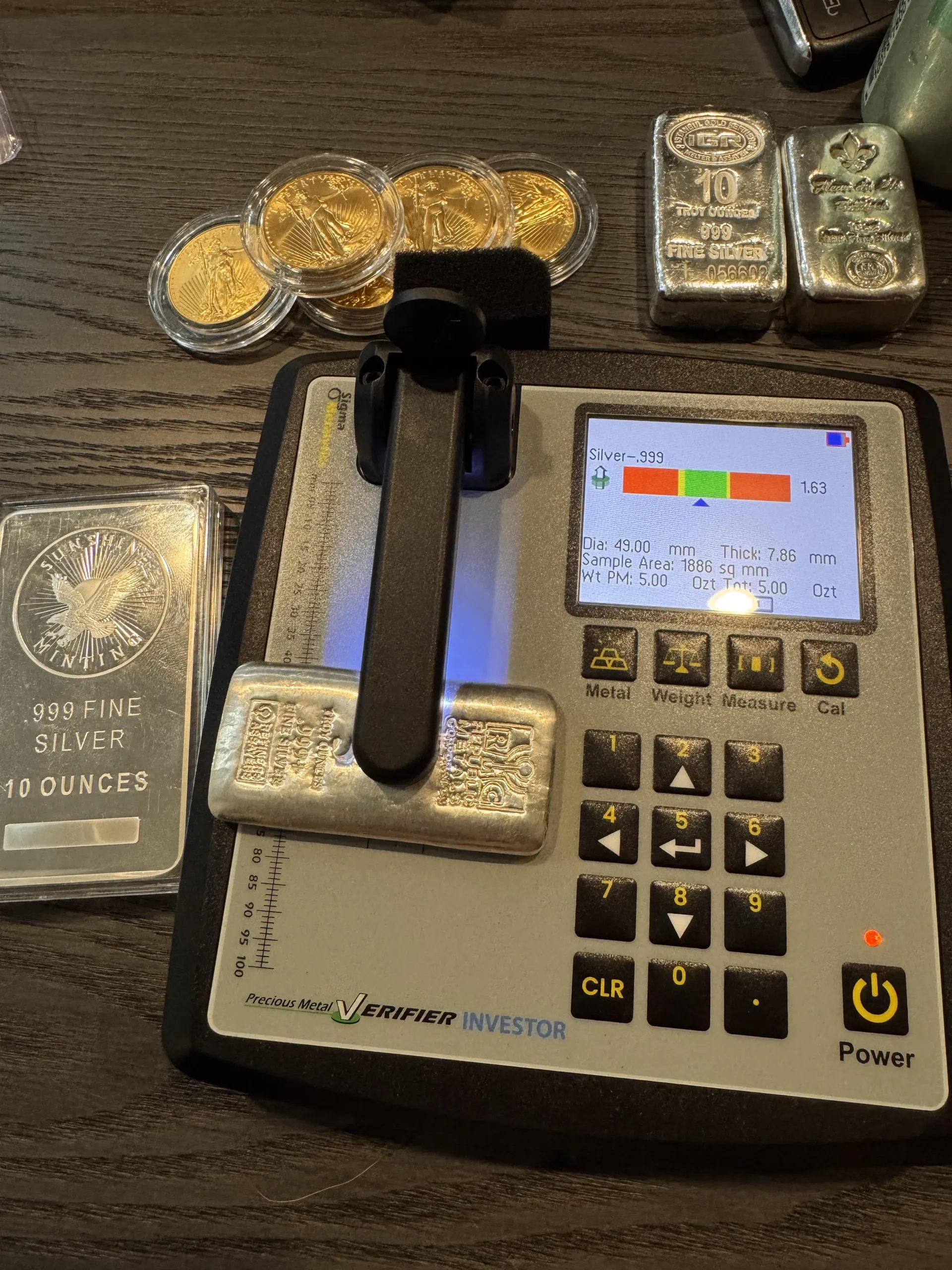 Silver bars and gold coins with a metal analyzer on a table.
