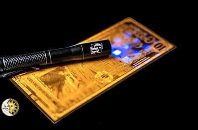 Black flashlight shining blue light on a gold-colored rectangular item against a black background.