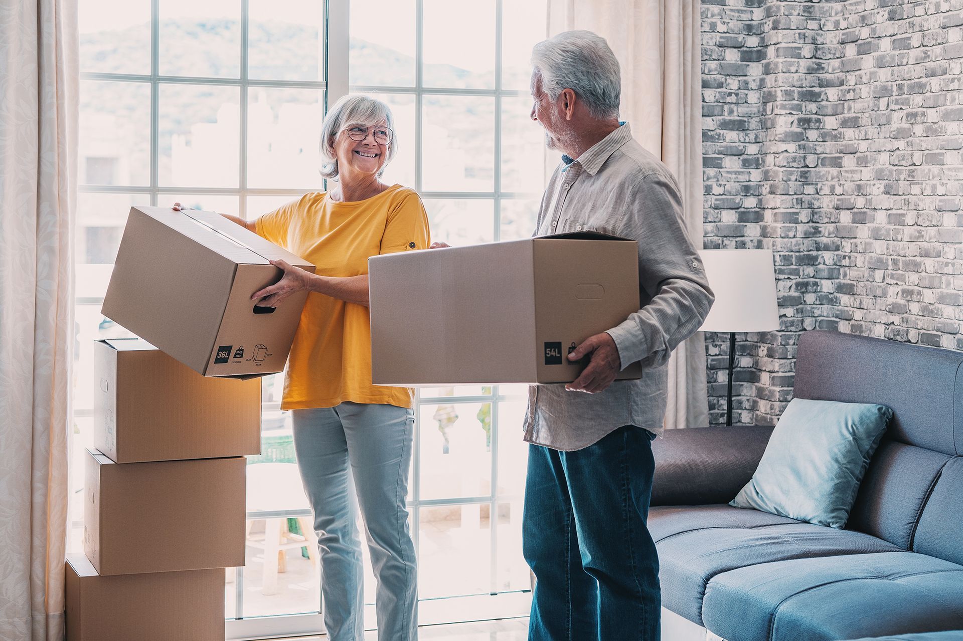 A man and a woman are carrying boxes in a living room.