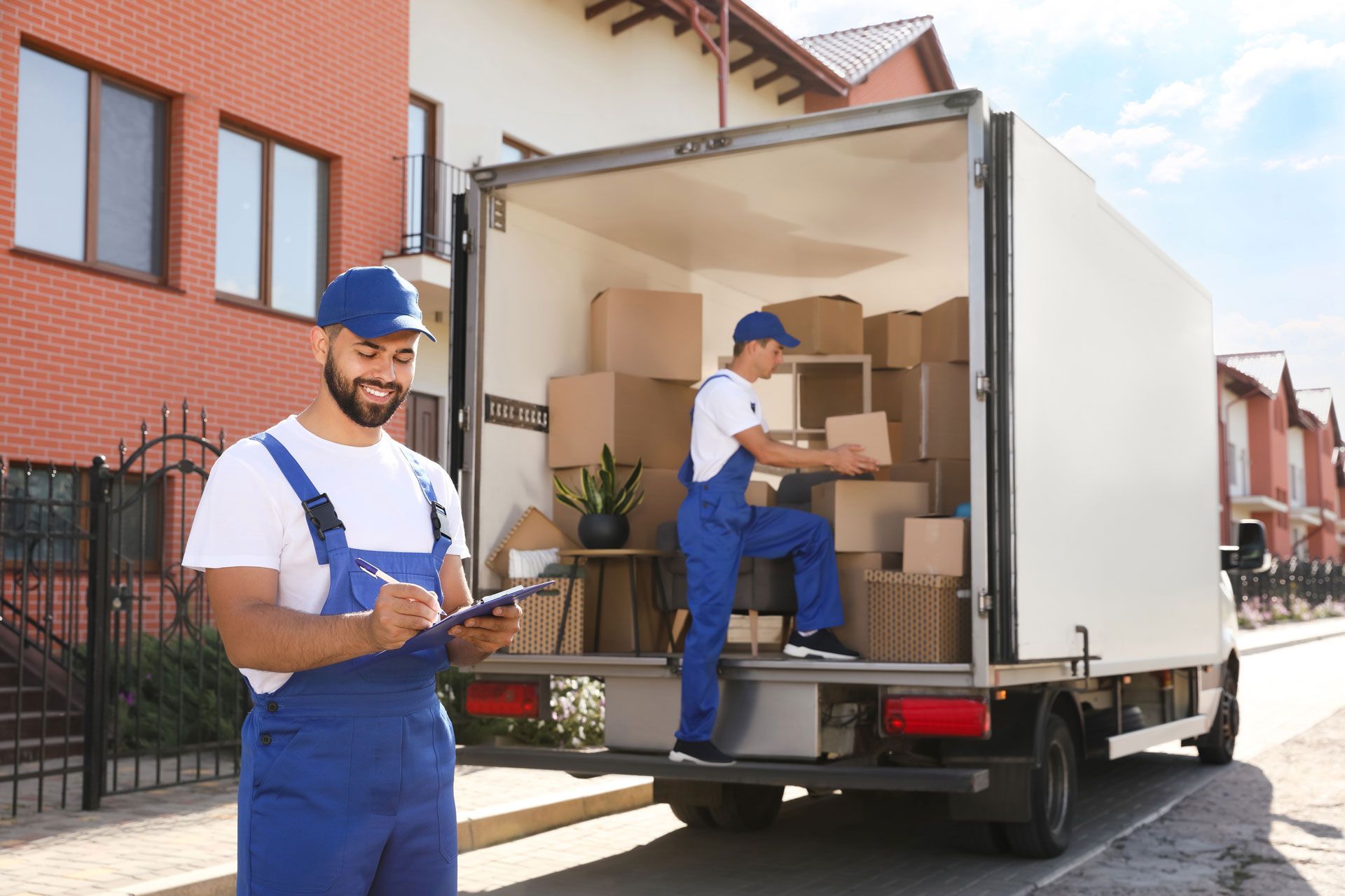 Two movers in blue uniforms and hats loading a truck with cardboard boxes on a sunny residential street.
