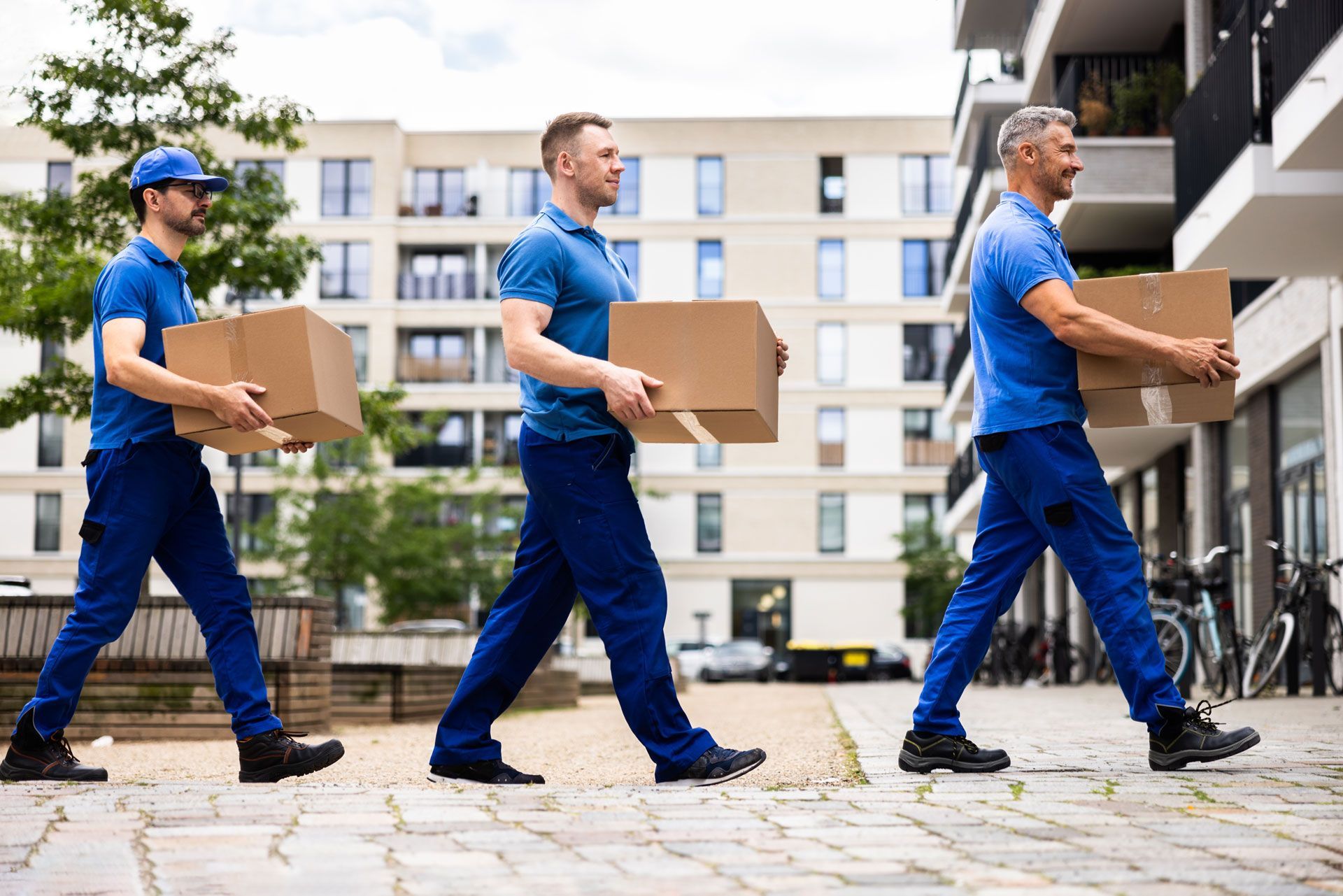 Three movers in blue uniforms walk in a line toward a residential building, each carrying a cardboard box.