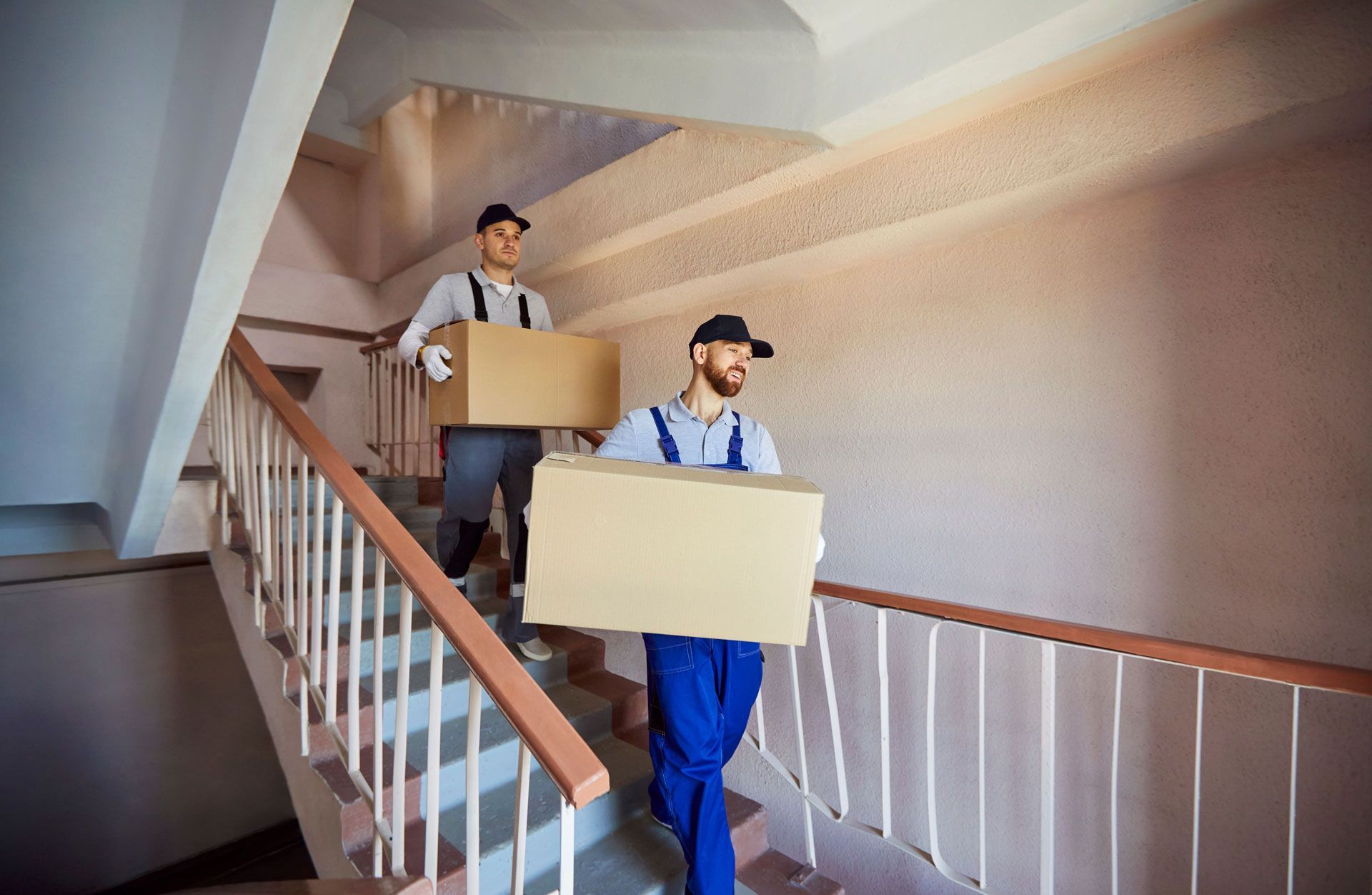 Two movers in uniform carry cardboard boxes down a staircase in a building.