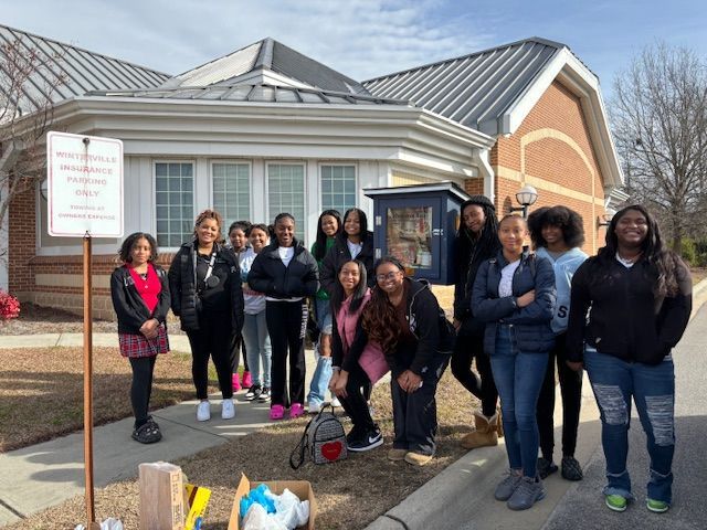 Group of girls stand in front of a building with a Little Free Library. Some hold bags, smiling.