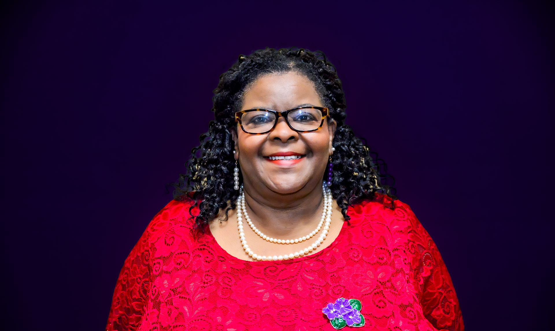 Woman in red lace top, wearing glasses and a pearl necklace, smiling; purple background.