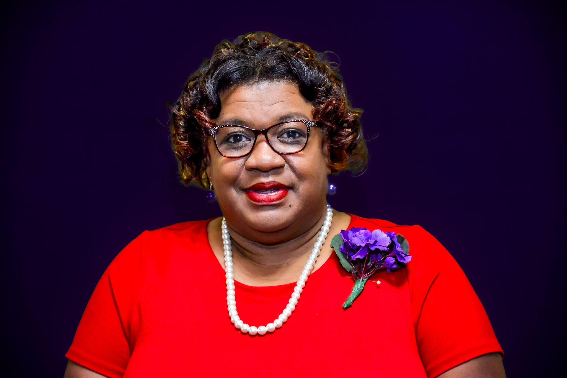 Woman with glasses and red top, smiling against a dark background.