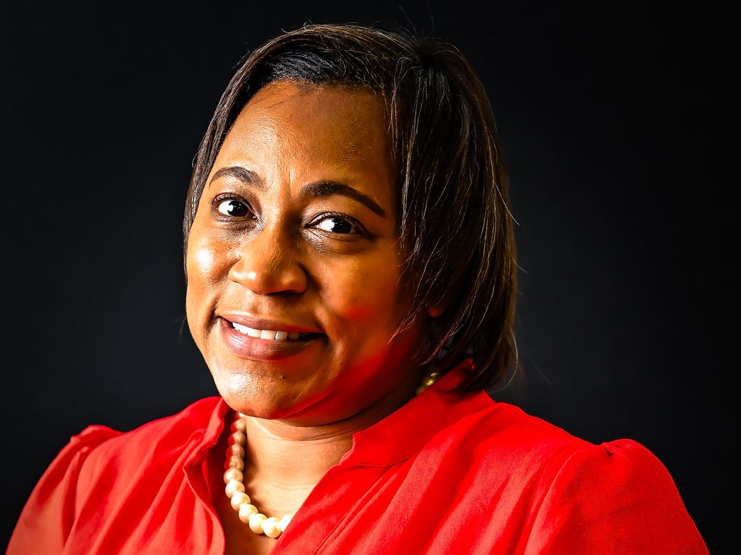 Woman smiling, wearing a red top and pearl necklace, dark background.