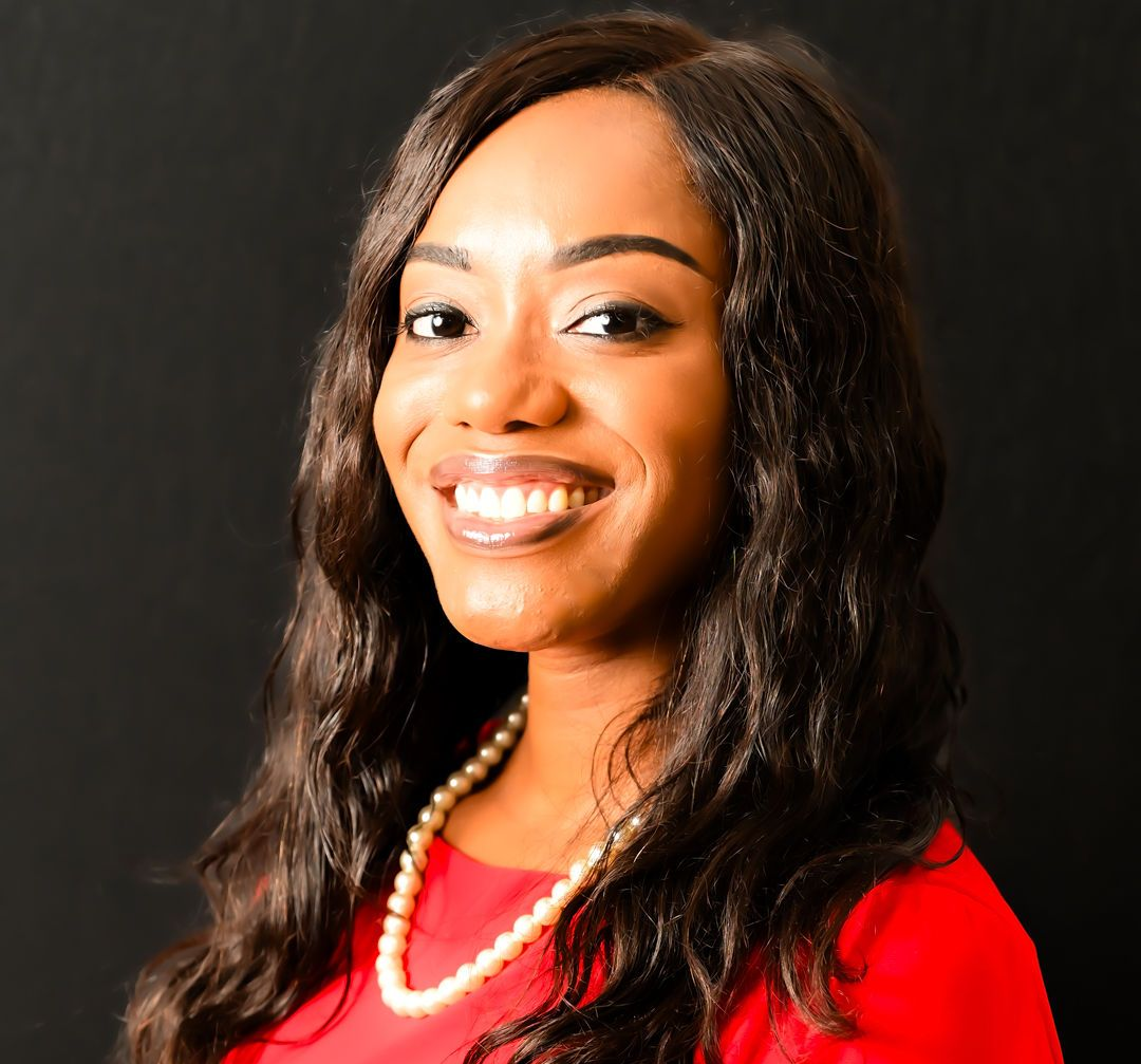 Woman with long dark hair, wearing a red dress and pearl necklace, smiling against a dark background.
