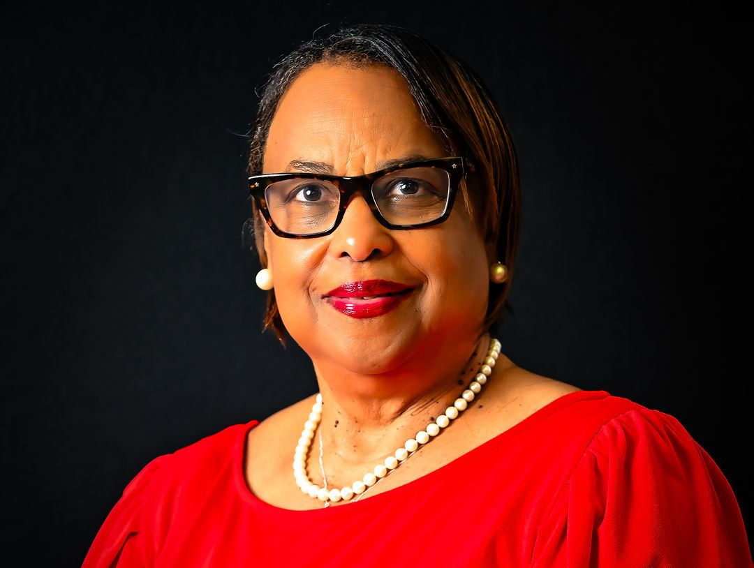 Woman in red dress and glasses, wearing a pearl necklace, smiles, with a black background.