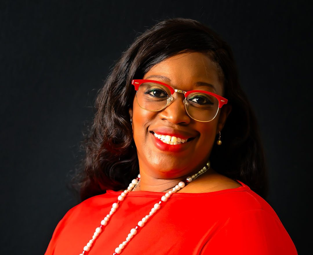 Woman with glasses and red dress smiling, wearing a pearl necklace and watch against a black backdrop.