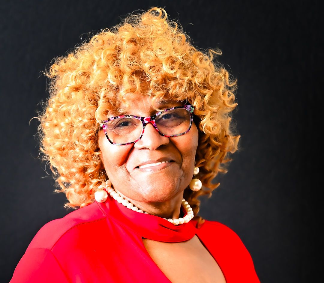 Woman with curly, blonde hair wearing glasses and a red dress, smiling against a black backdrop.