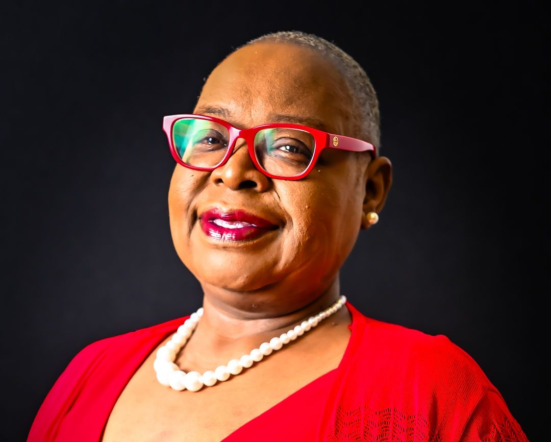 Woman with a shaved head wearing red glasses, top, and jacket smiles, with pearls, against a black background.