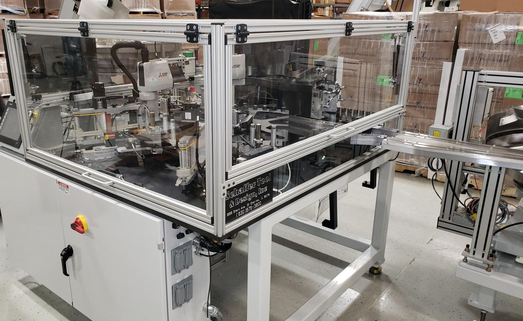 An industrial automated assembly machine with a clear safety enclosure, standing on a white base in a warehouse.