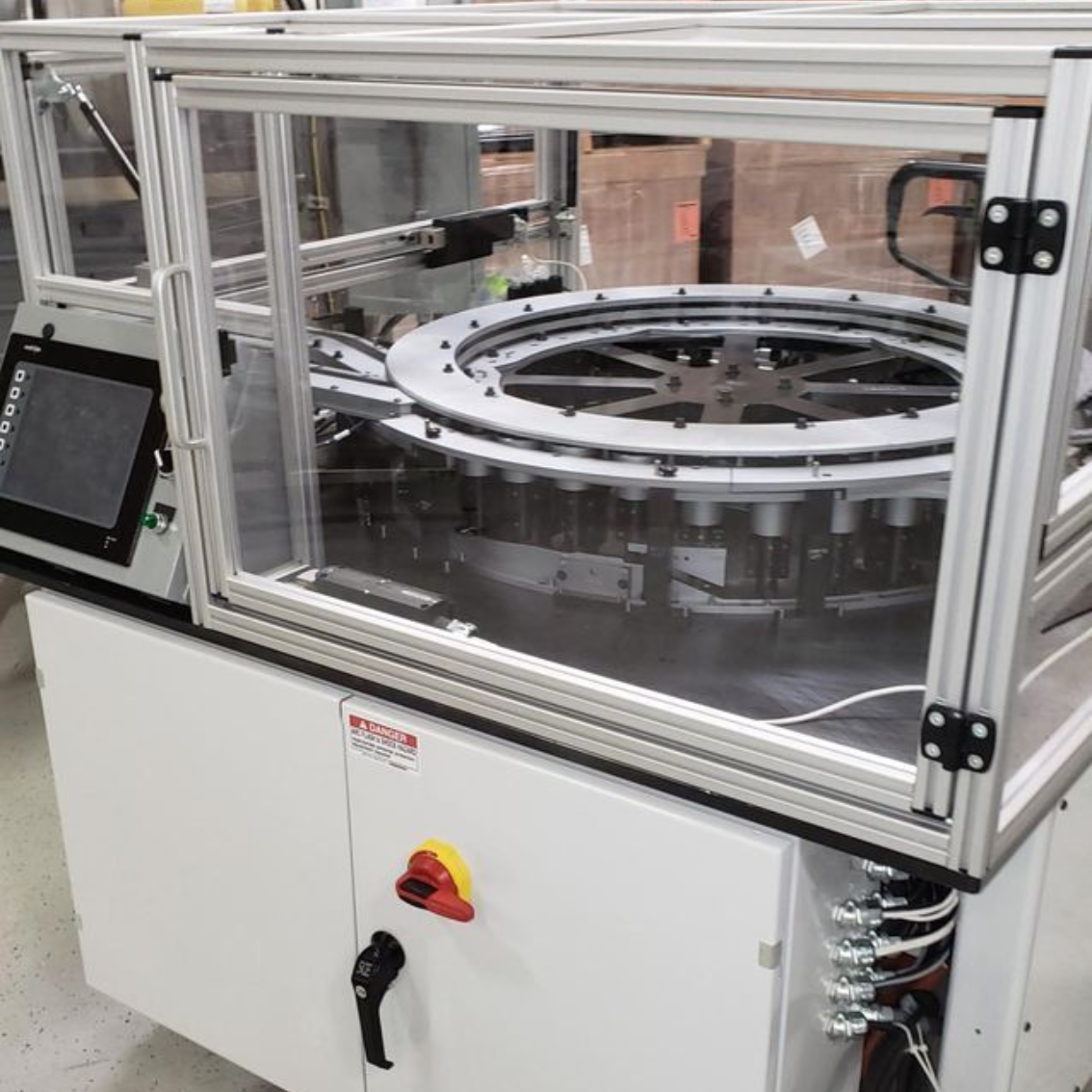 An industrial automated assembly machine with a clear safety enclosure, standing on a white base in a warehouse.