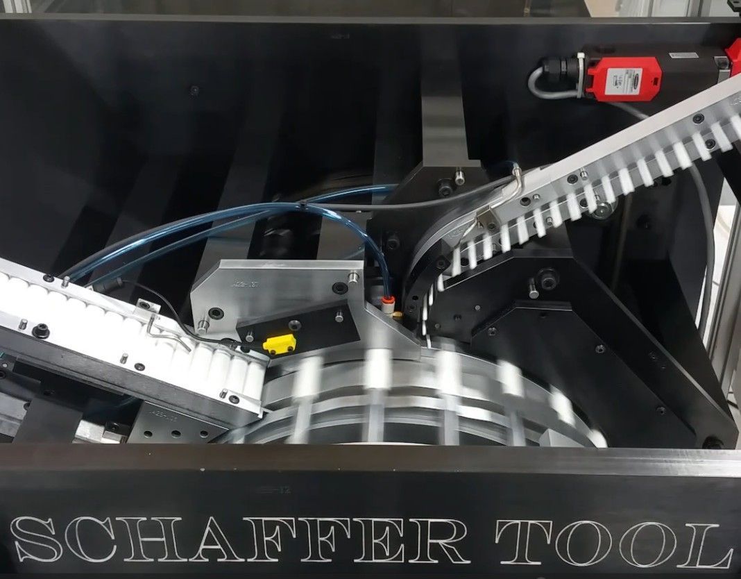 An industrial automated assembly machine with a clear safety enclosure, standing on a white base in a warehouse.