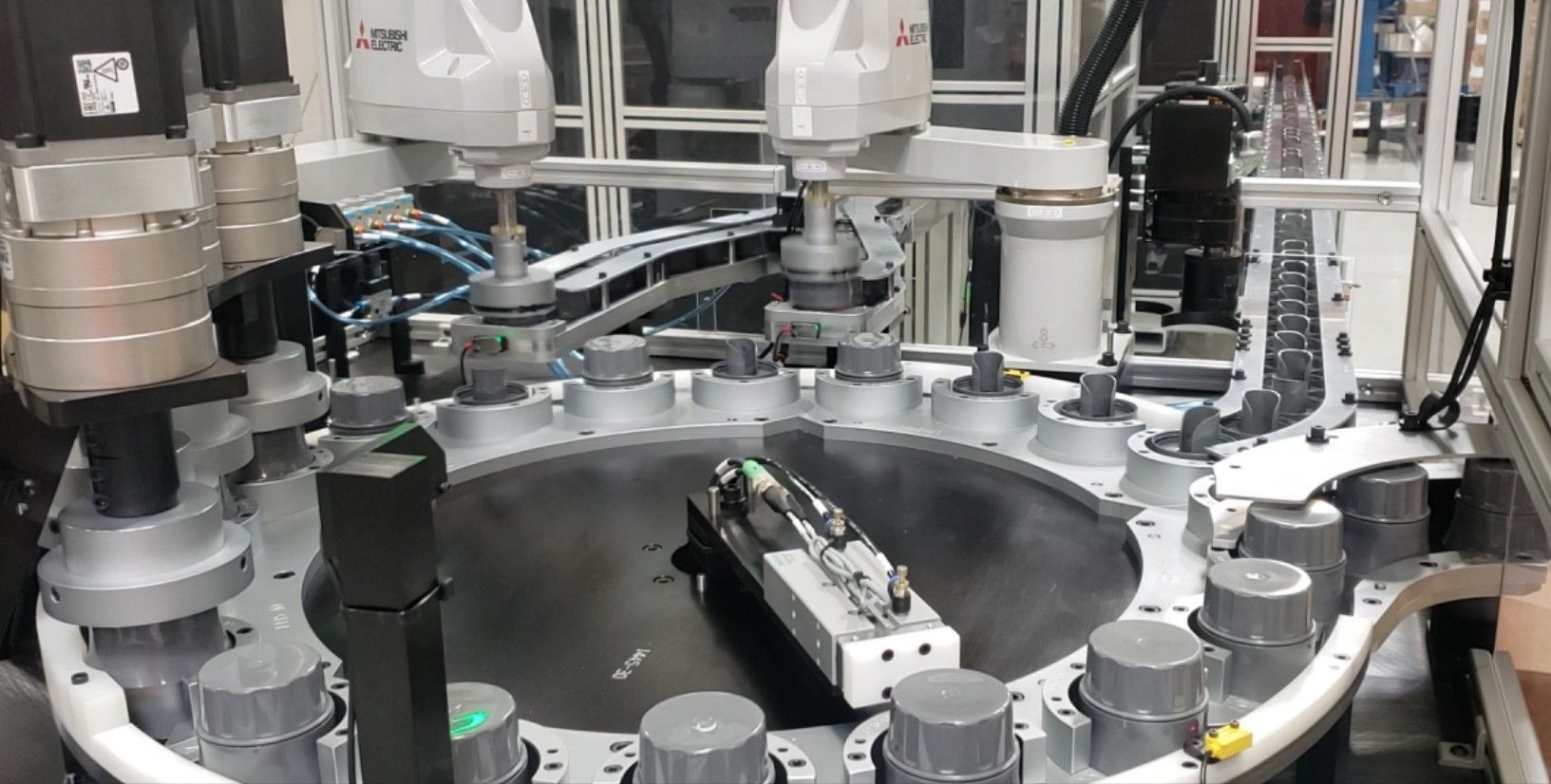 An industrial automated assembly machine with a clear safety enclosure, standing on a white base in a warehouse.