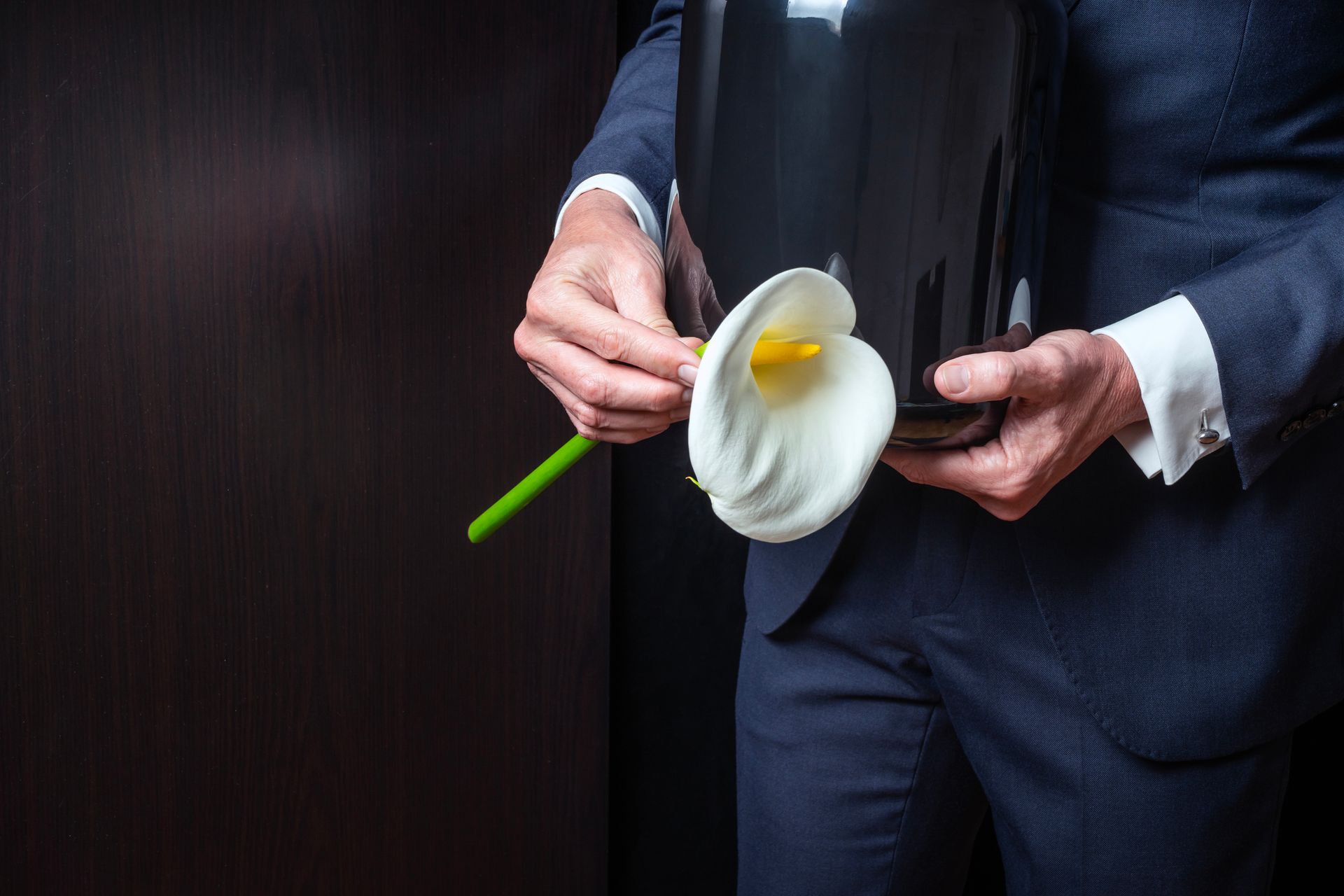 Elegant mourning man holds cremation urn and white calla lily at funeral service.