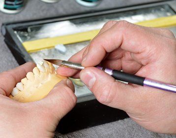 Hands using a brush to apply substance to a set of ceramic teeth in a lab setting.