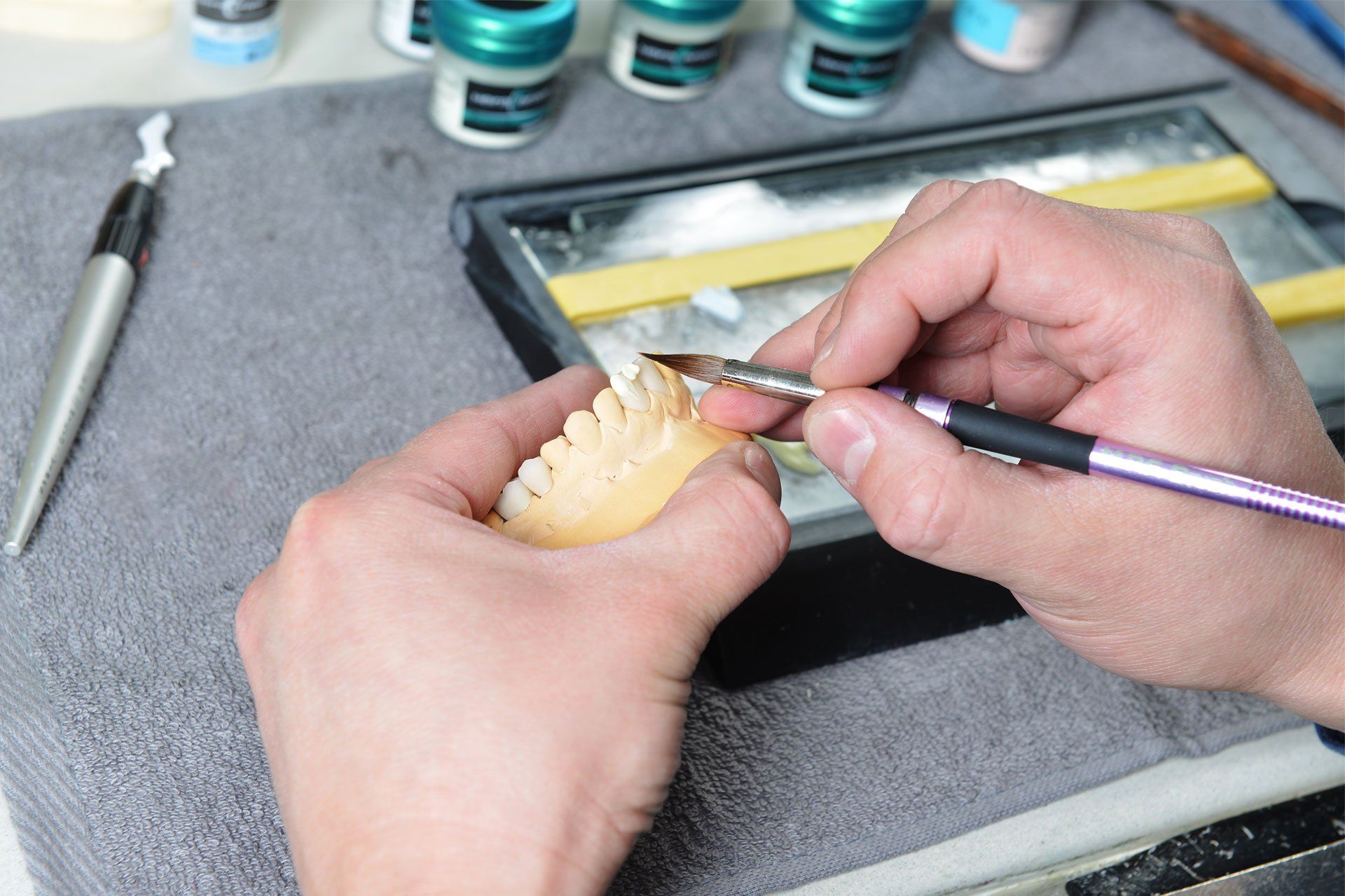 Hands applying color to a dental model with a brush. Tools and paint jars visible.