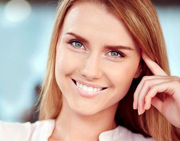 Blonde woman with blue eyes smiles, hand to head, in an office setting.