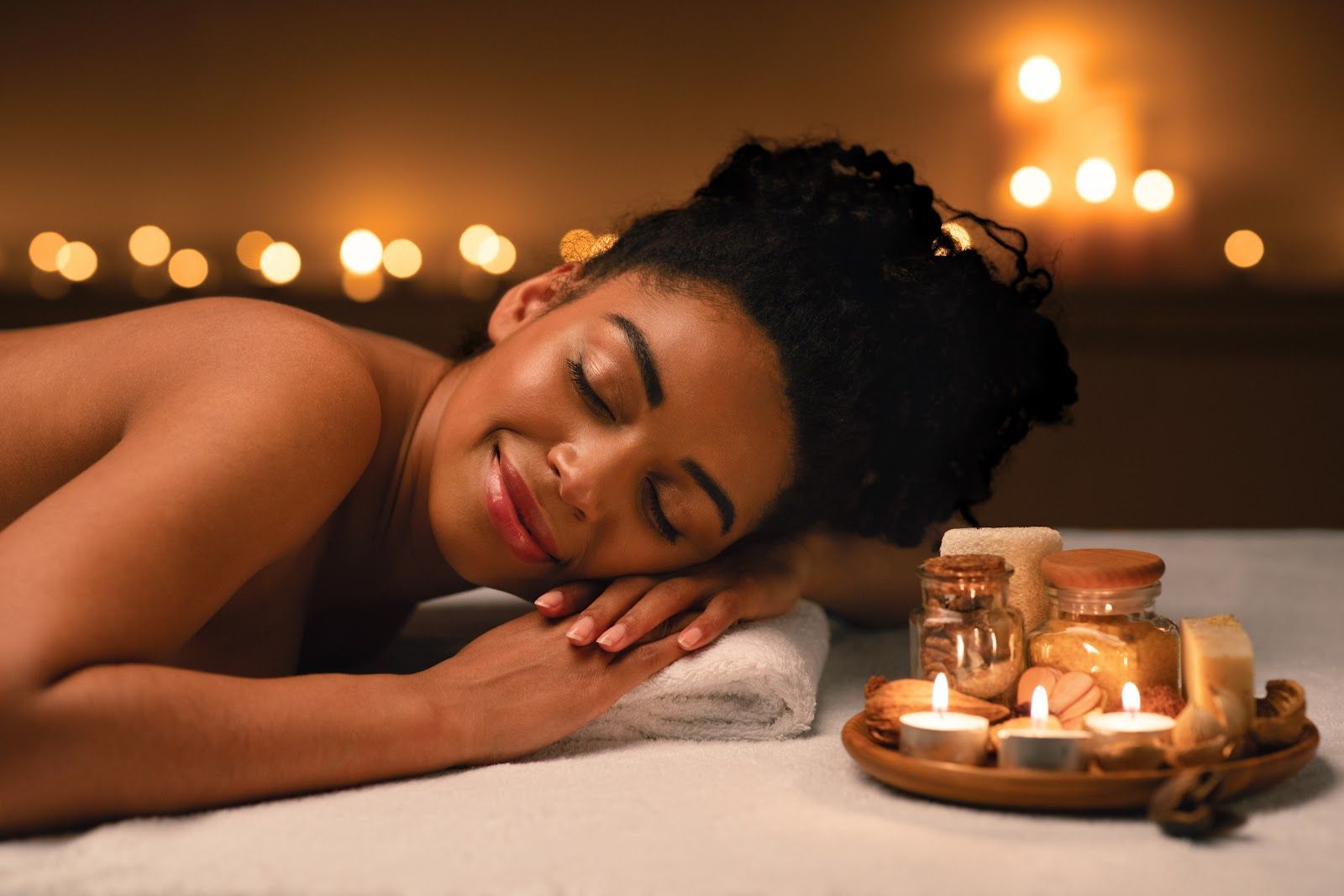 Woman lying on massage table, eyes closed, smiling. Candles and spa products in the background.