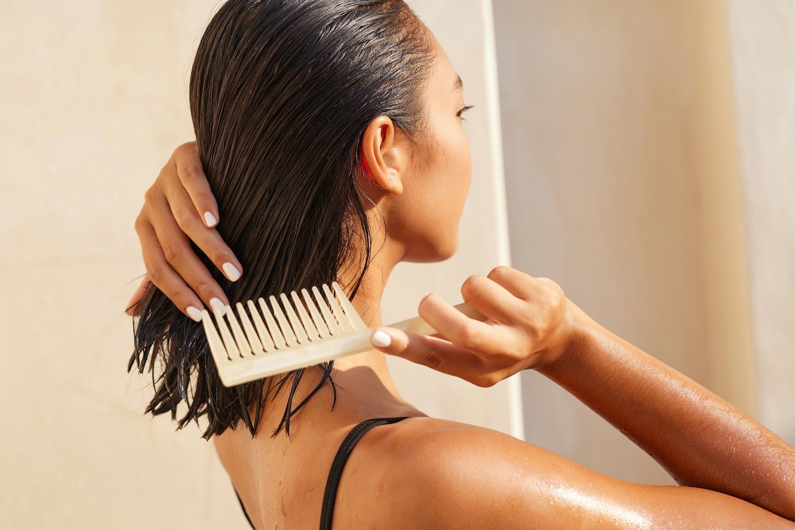 Woman combing wet hair in a shower, back view; pale comb; sunlight.