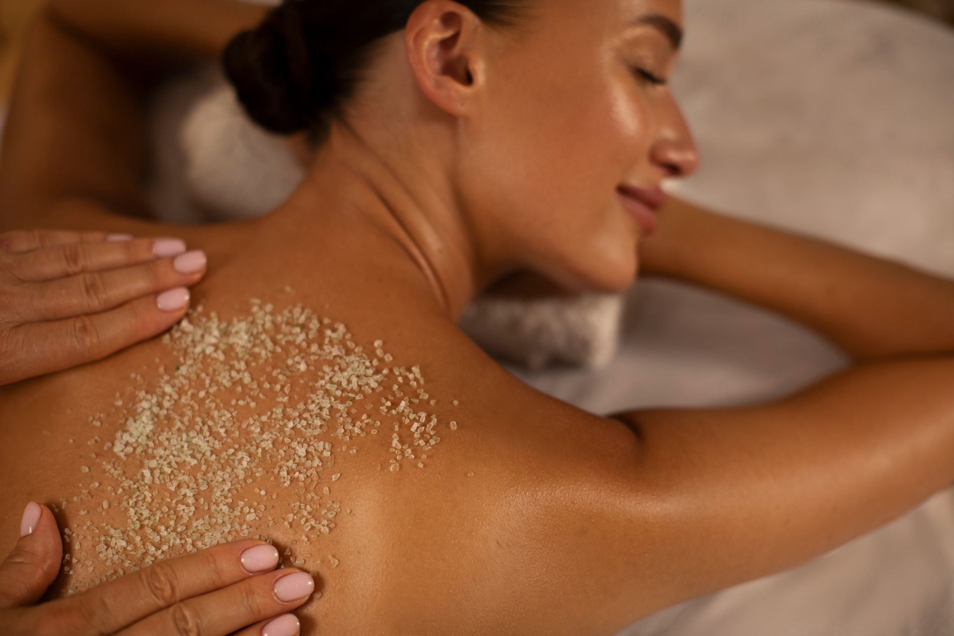 Woman receiving a back scrub at a spa. Hands apply granular product to skin, which is smooth and glowing.