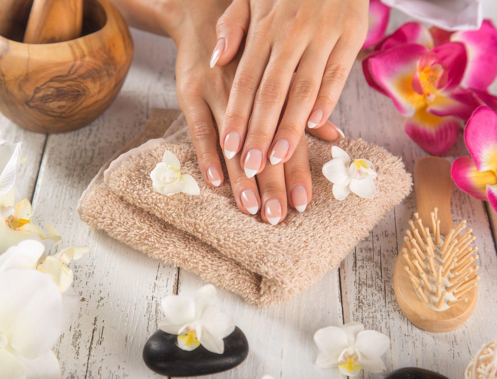 Hands with French manicure rest on a folded towel with flowers. Spa setting with wooden items.