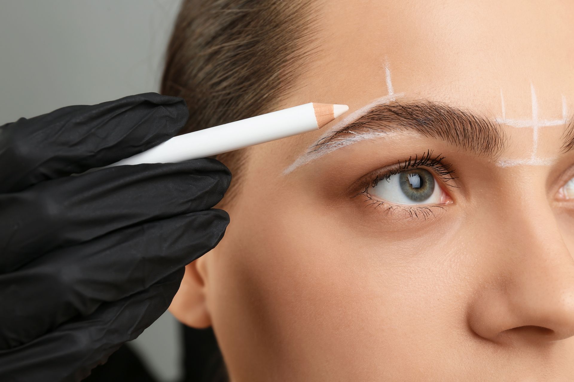 Gloved hand using a white pencil to mark a woman’s eyebrow for cosmetic treatment