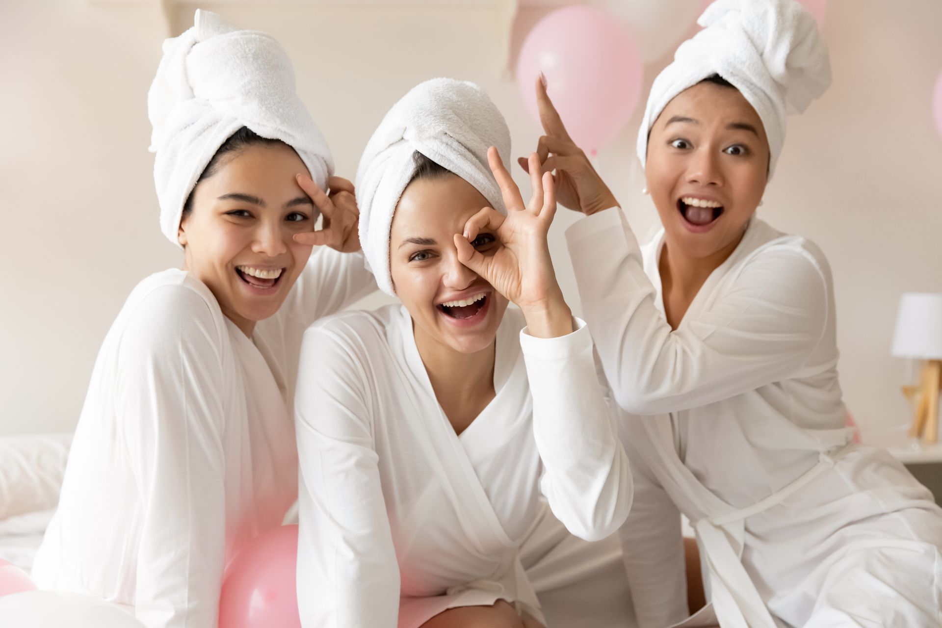 Three people in white robes and towels smiling together with pink balloons in a bright room