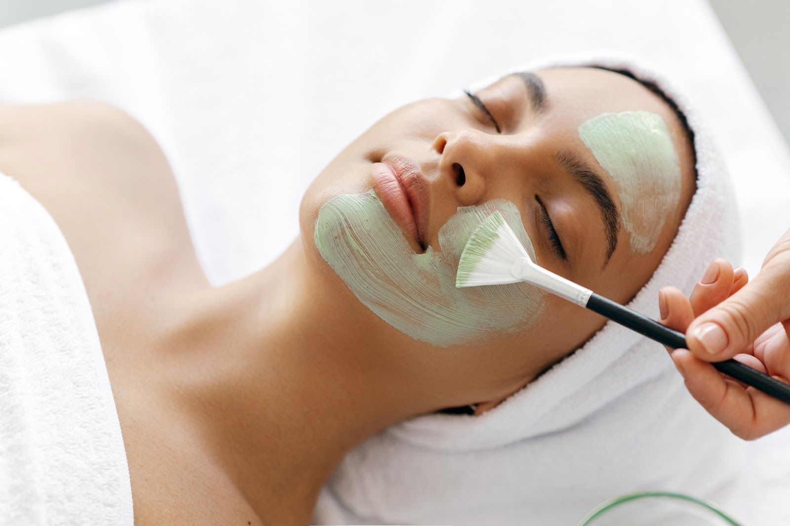 Woman with facial mask, being treated with a brush in a spa setting.