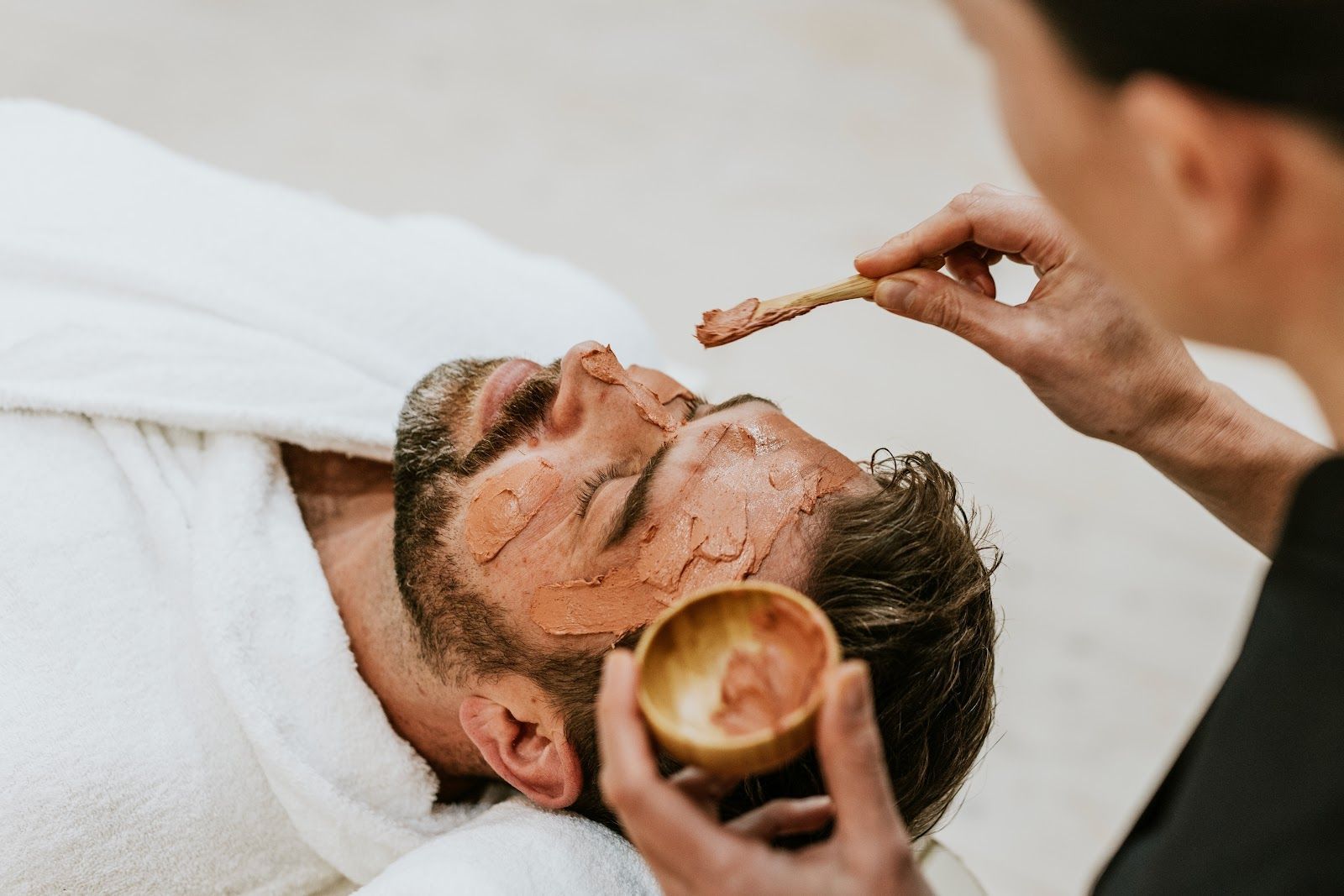 Person receiving facial treatment, therapist applying mask with small wooden spatula.