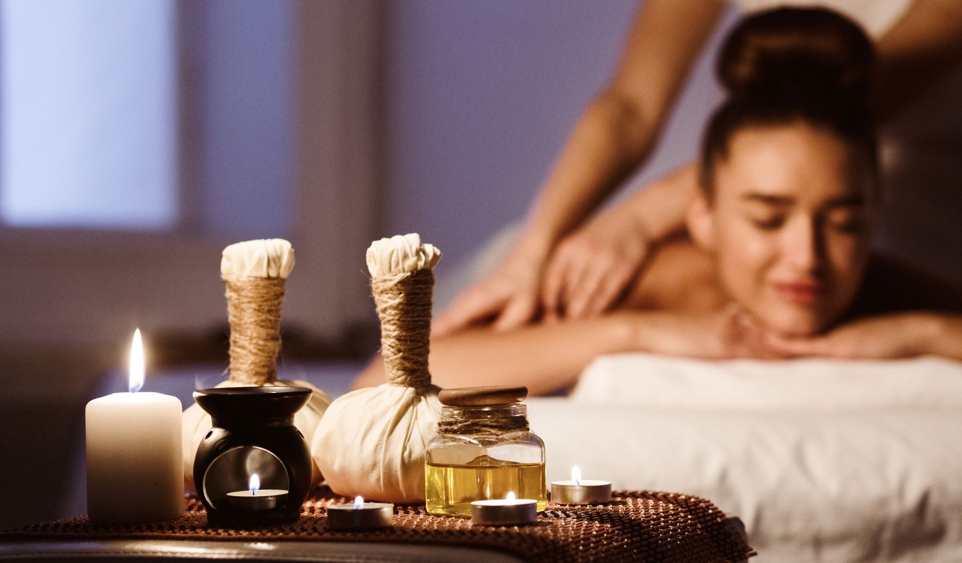 Woman receiving back massage at spa; candles, herbal bundles, and oils in foreground.