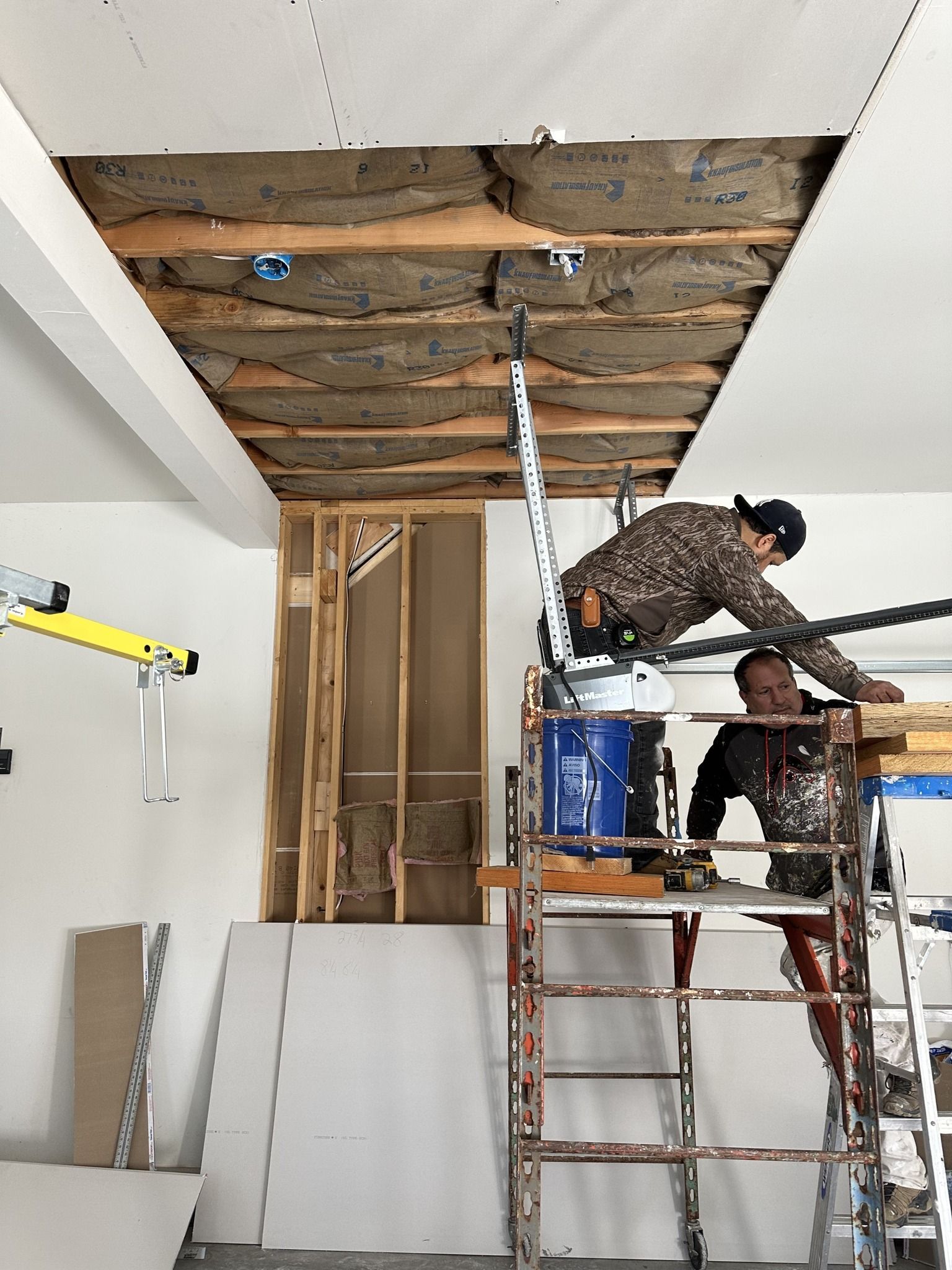 Workers repairing a ceiling on scaffolding in a room with exposed beams and drywall panels.